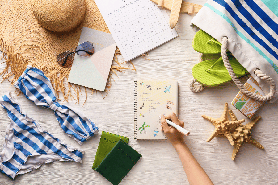 Flat lay of a table filled with beach day and travel accessories. A woman is writing her travel checklist.